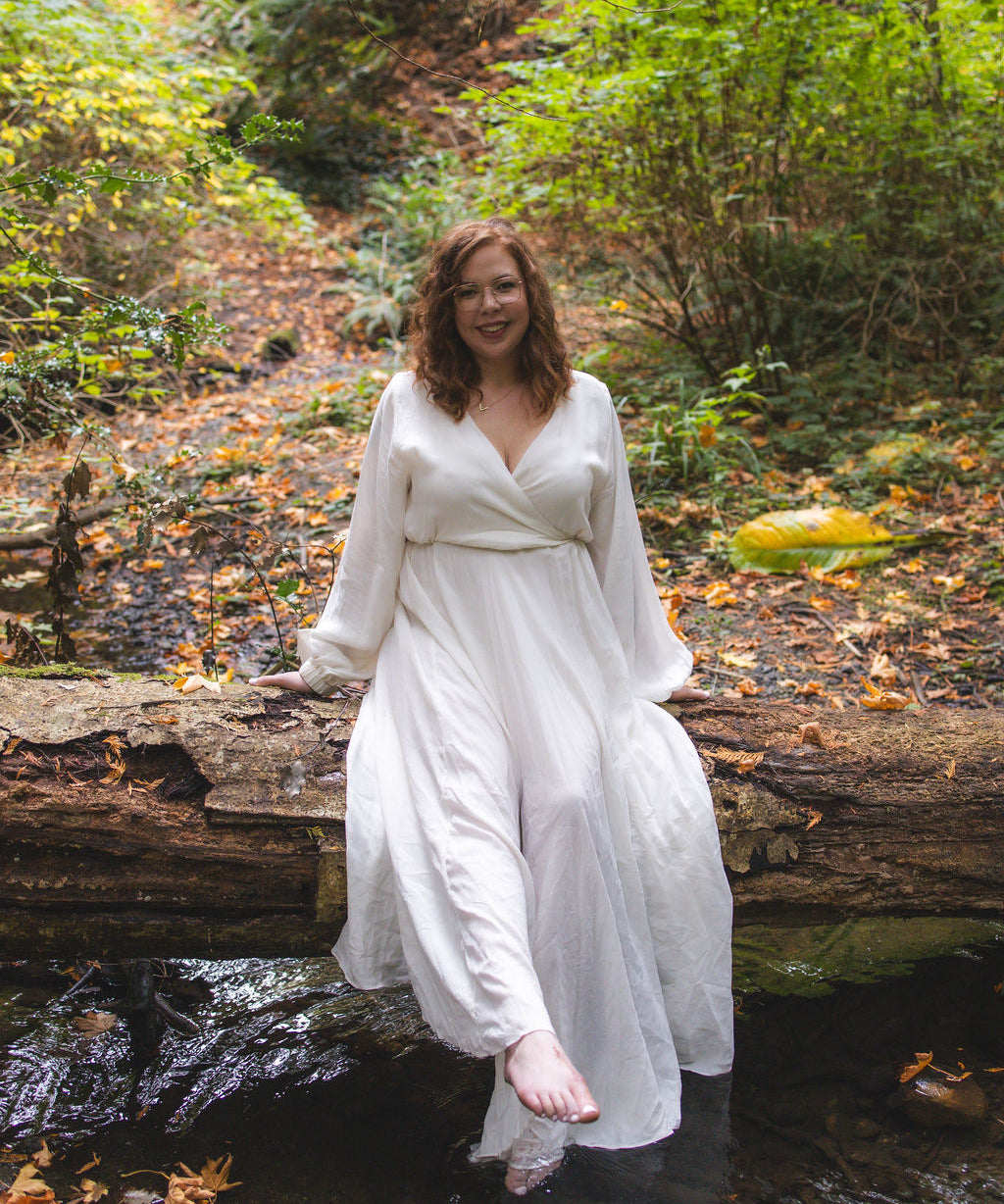 Taylor, WAASEYA founder in a white dress standing on a log over a stream in a forest