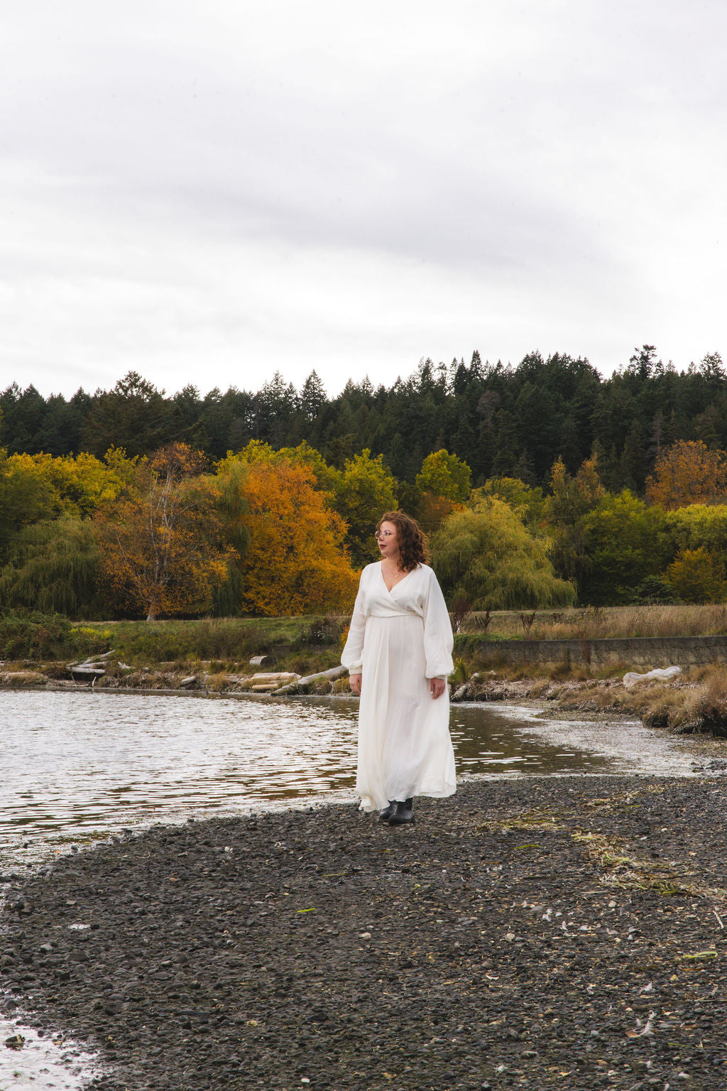 Waaseya Founder, Taylor LaVallee, in a white dress, standing on a rocky shoreline with trees and water in the background